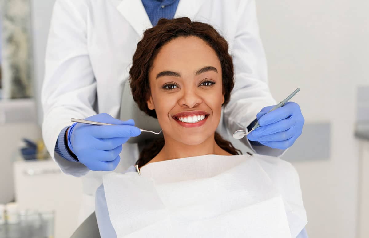 smiling woman in dental chair with dentist holding tools behind her